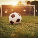 A football sits on a grass pitch while the sun sets in the background of a goal.