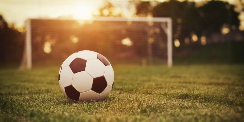 A football sits on a grass pitch while the sun sets in the background of a goal.